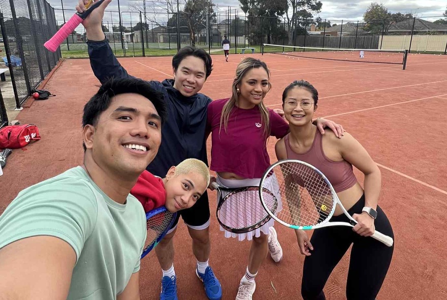 A group of smiling people standing on a tennis court, some holding racquets, taking a selfie.