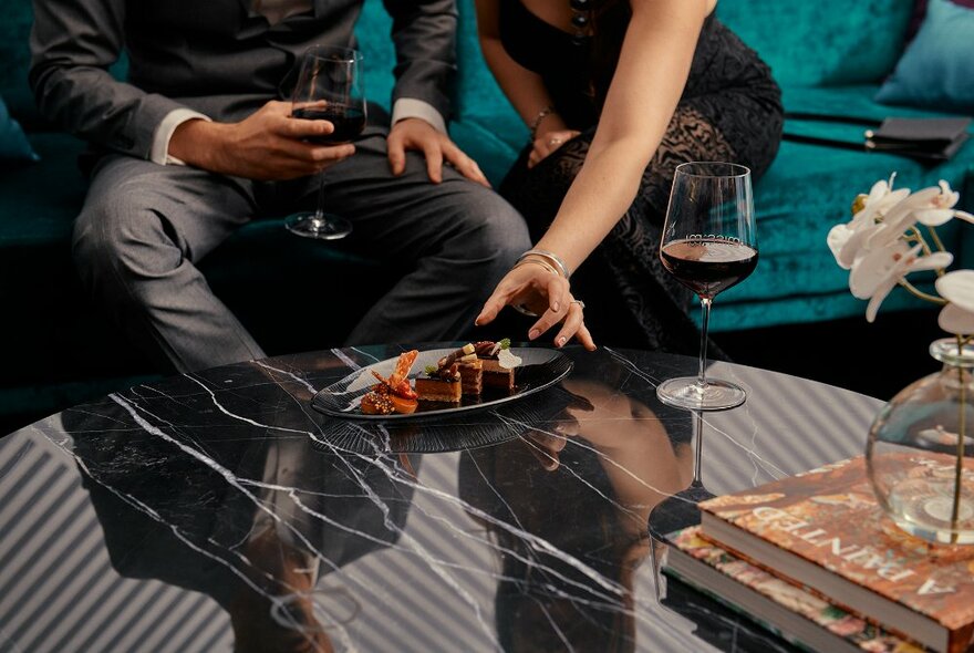 Two people seated on a couch and reaching in to select food from a plate that is on a coffee table in front of them, a glass of wine nearby.