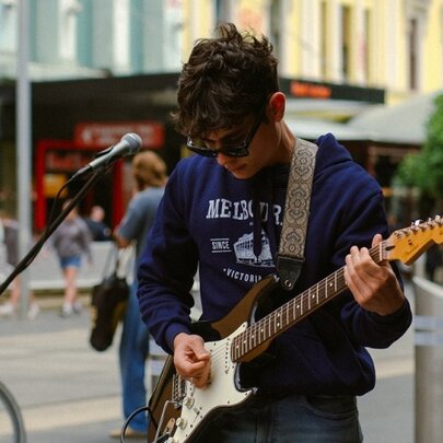 A busker performing in the street playing an electric guitar with his head facing downwards looking at the instrument. 