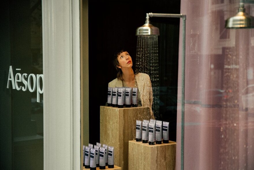 Person standing at an installation inside an Aesop window, looking up at a shower head that has droplets of water coming out if it.