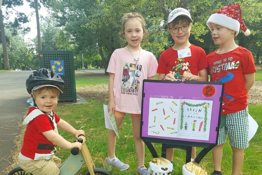 Four children in a park, standing behind a picture book, one of them wearing a Christmas hat.