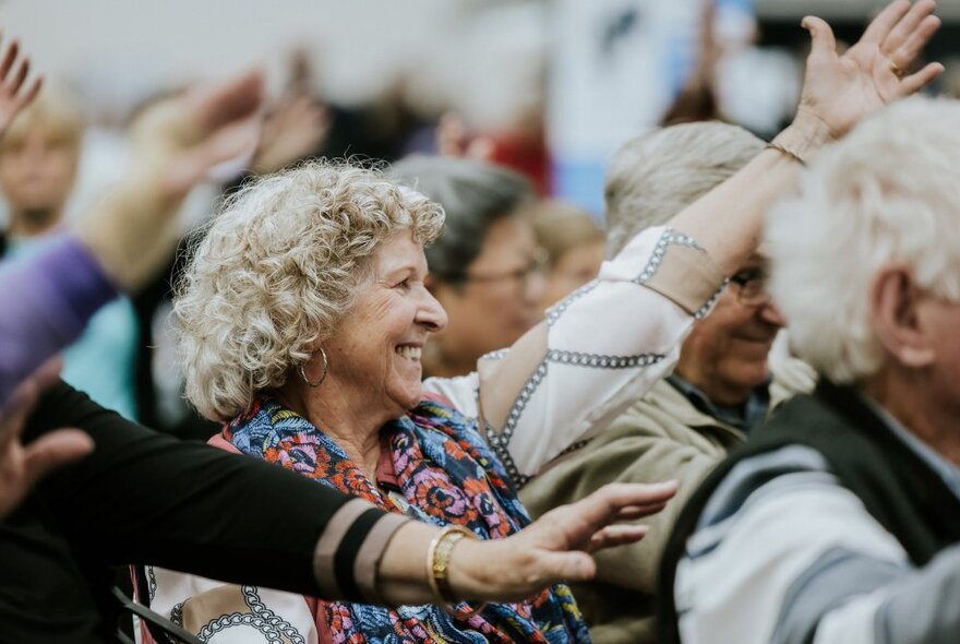 Audience members at an expo, seated in chairs, holding one arm up above their heads and stretching.