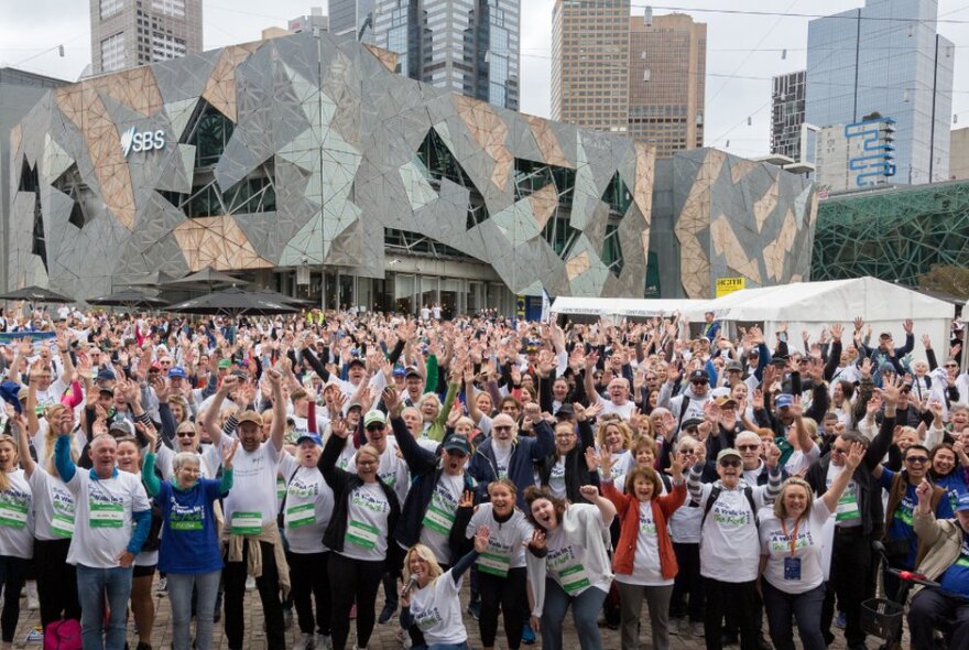 A large crowd of people wearing charity t-shirts at Fed Square, each waving their arms in the air.