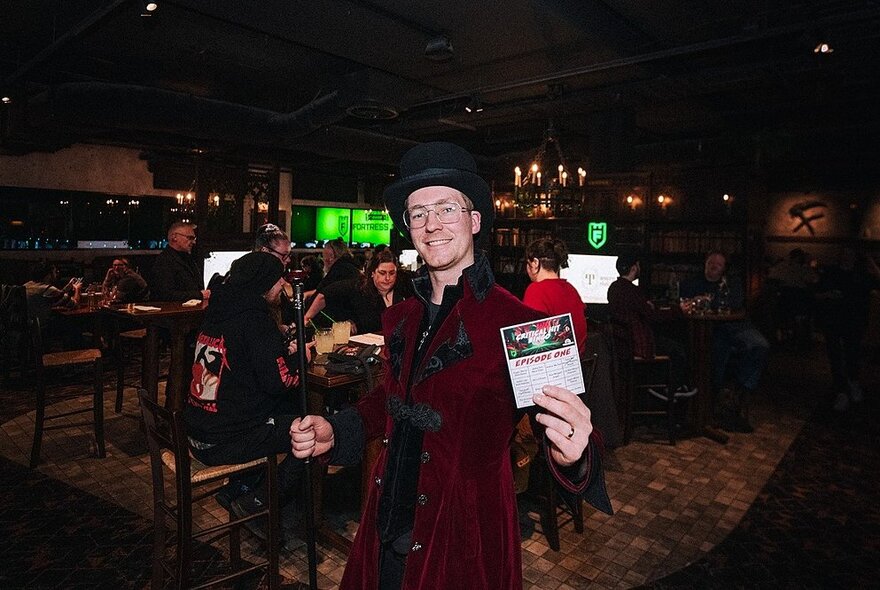 Man with hat and glasses standing in a dark bar, holding a card.
