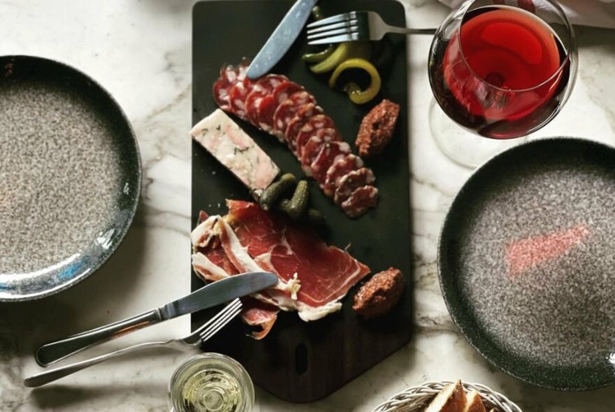 Charcuterie on a platter with plates, cutlery and glasses of wine.