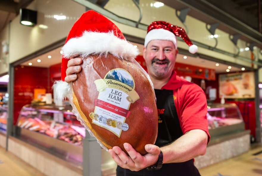 A market stallholder wearing a Santa hat, holding a ham wearing a Santa hat.