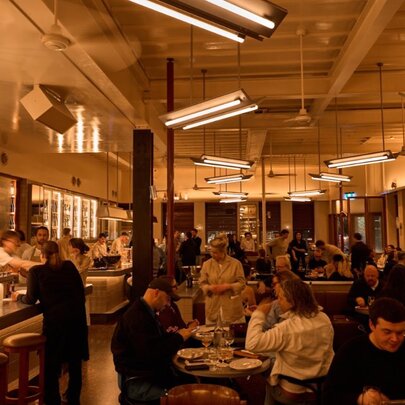 Warmly lit restaurant interior with bench and table seating, open kitchen and overnight strip lights.