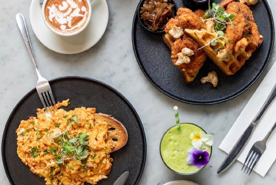 Scrambled eggs on toast and breakfast waffles on two black plates seen from above, alongside a green smoothie and a cappucino.