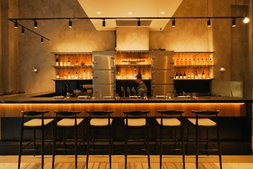 Six high bar chairs facing a modern service area, with backlit shelves of bottle of alcohol and large stainless steel fridges on the rear wall.