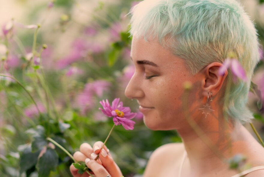 Outdoors in a field, a side profile of a person's head, their eyes are closed and they are holding a pink flower to their nose.