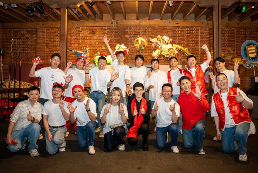 The True Colours male choir in white t-shirts and blue jeans, casually posing in an indoor venue, with arms and hands raised in friendly thumbs up and waving gestures.