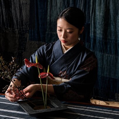 Traditional Japanese flower arrangement, or Ikebana, being created by a young woman wearing a kimono on a table inside a retail or gallery space.