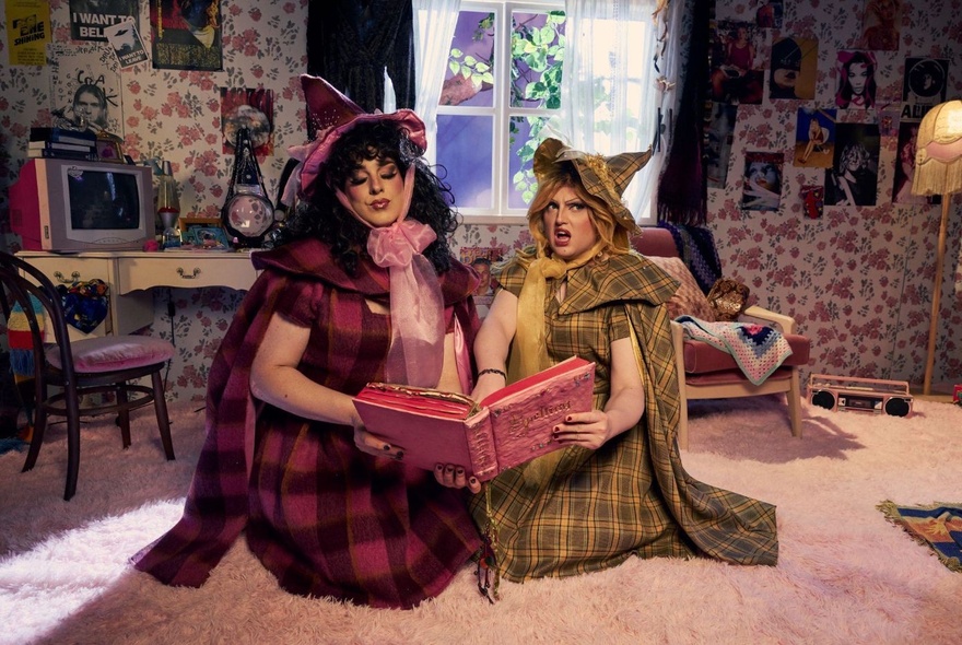 Two performers in drag, wearing plaid witch outfits and hats sit on a rug, reading a large red book together in a cluttered room.