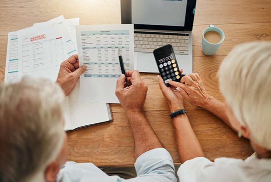 An older couple with a smart phone and laptop, looking at printed documents.