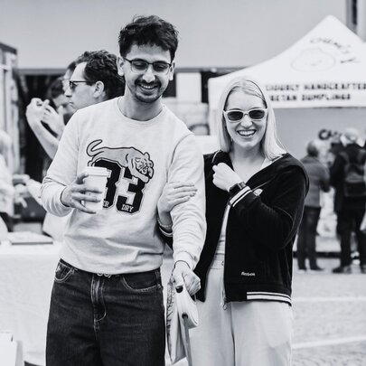 Black and white photograph of a young couple at an open air market, small marquees in the background.