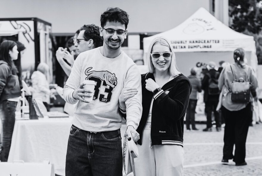 Black and white photograph of a young couple at an open air market, small marquees in the background.