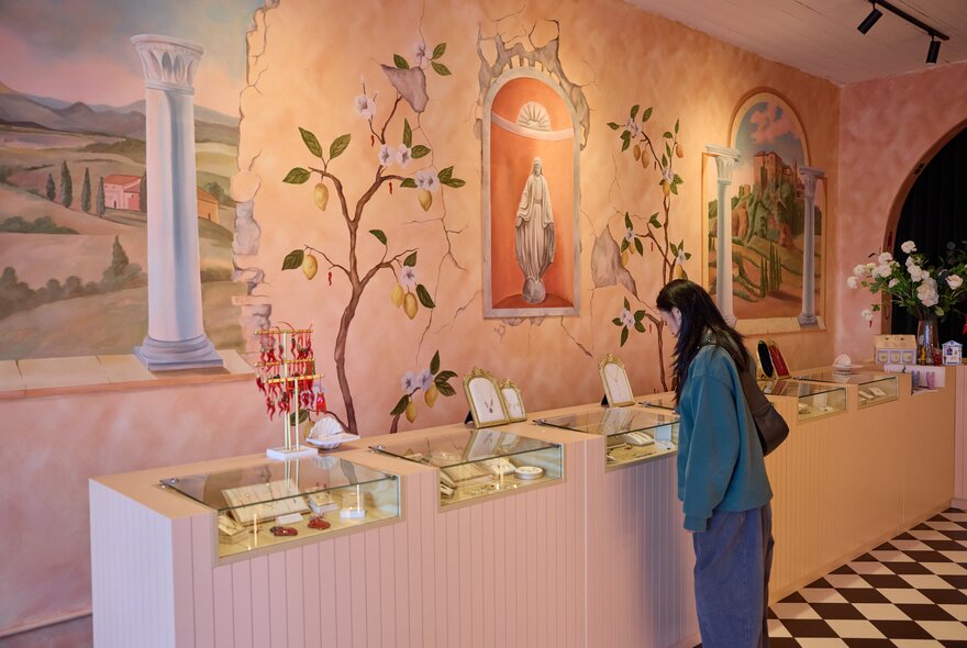A woman looking at glass cases of jewellery in a store with a pink mural of lemon trees and Italian scenery on the wall.
