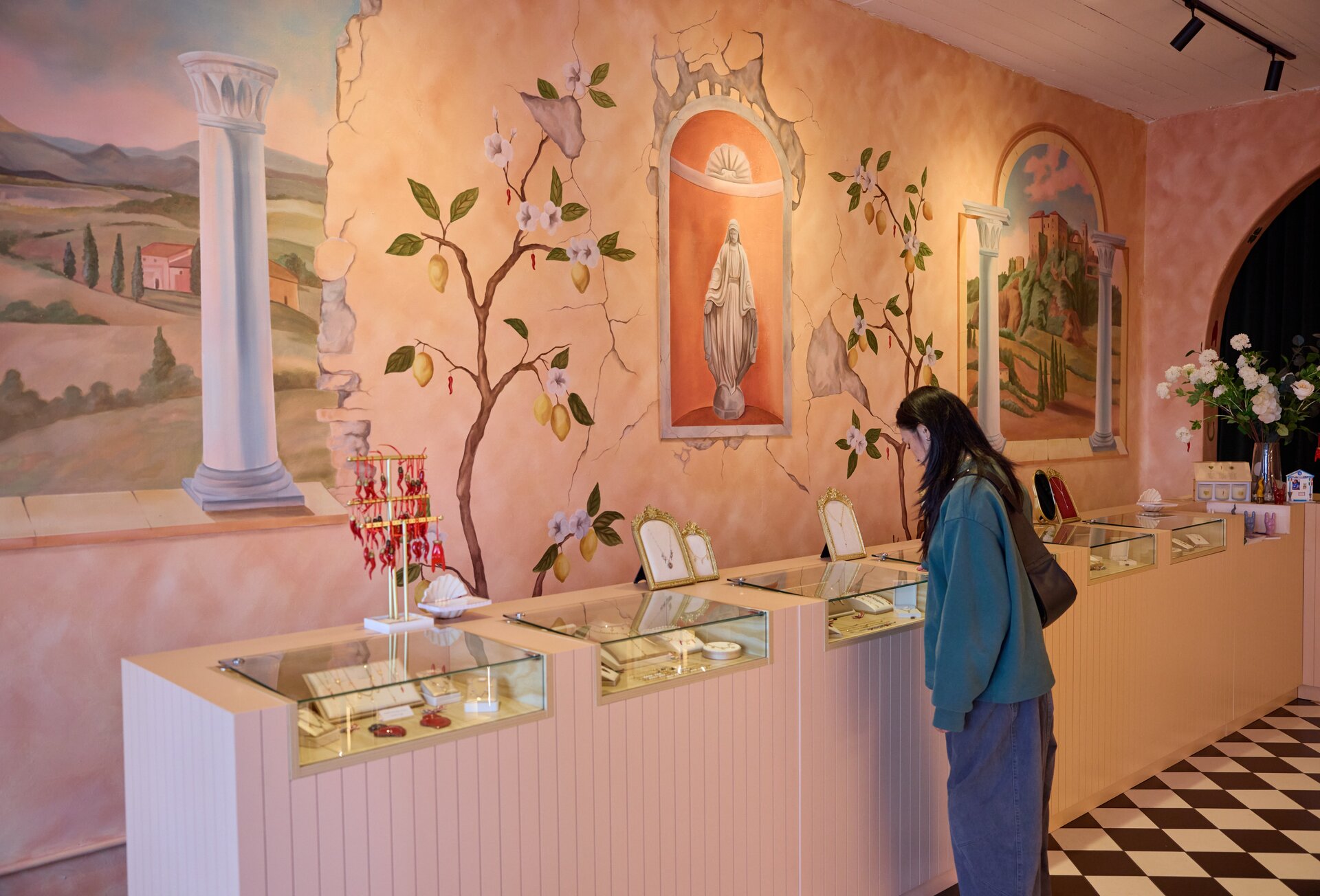 A woman looking at glass cases of jewellery in a store with a pink mural of lemon trees and Italian scenery on the wall.