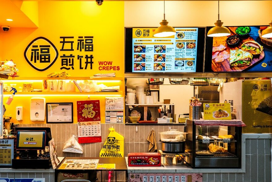 The exterior of the Wow Crepes food kiosk at Kmart Centre food court, showing a small space and service counter, with signs and menu items displayed on the rear wall, and kitchen and shop paraphernalia.