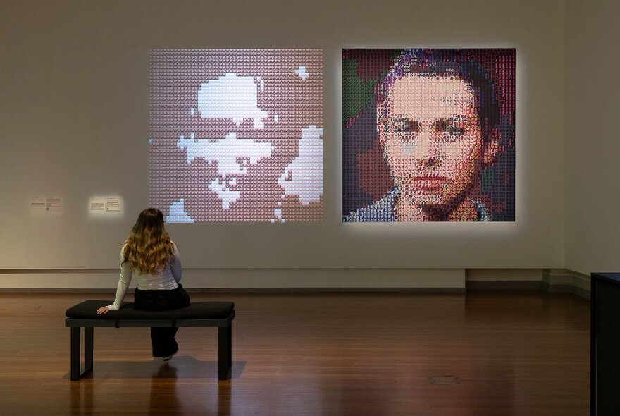 A gallery space with timber floors and white walls, with a projection of two pixellated faces on the wall, one more abstract than the other, and a patron seated on a wooden bench.