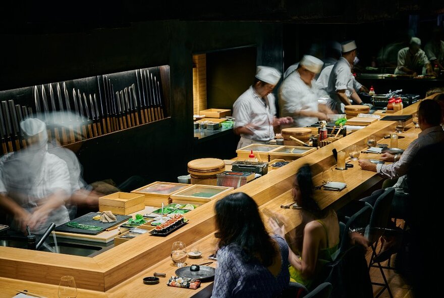 Several chefs preparing Japanese food in front of dining patrons who are seated at a high wooden bar in front of them.