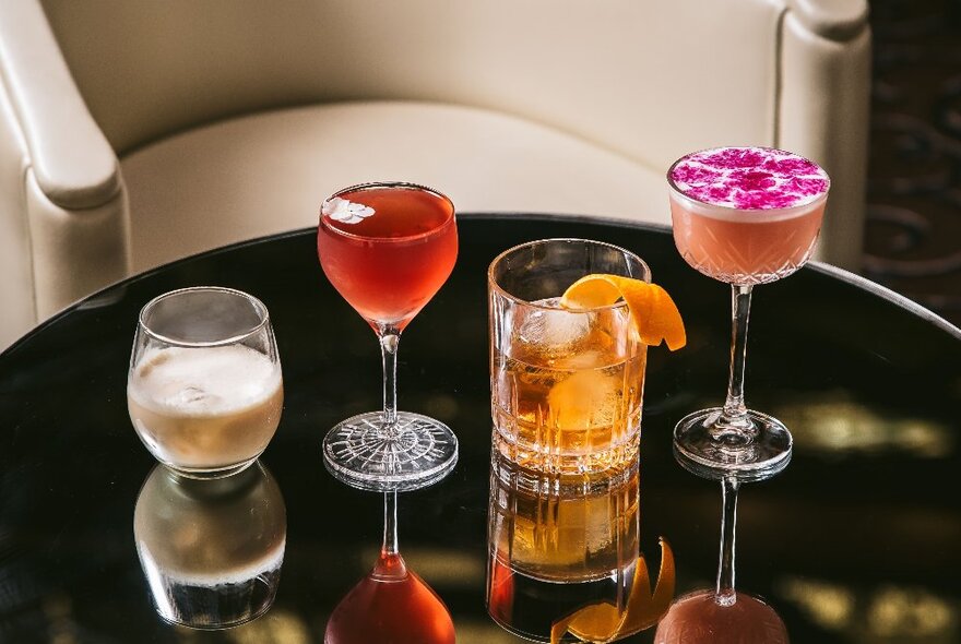 Four cocktails in different shaped glasses on a reflective tabletop, with a club armchair in the background.