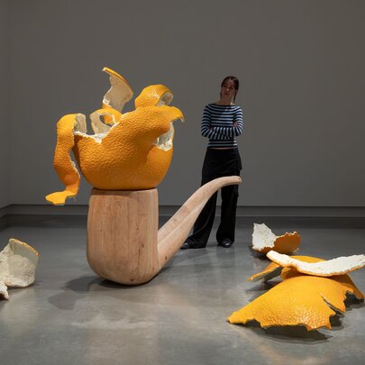 A person in a gallery setting looking at a sculpture of an oversize wooden pipe with a sculpture of a giant peeled mandarin balanced on the pipe.