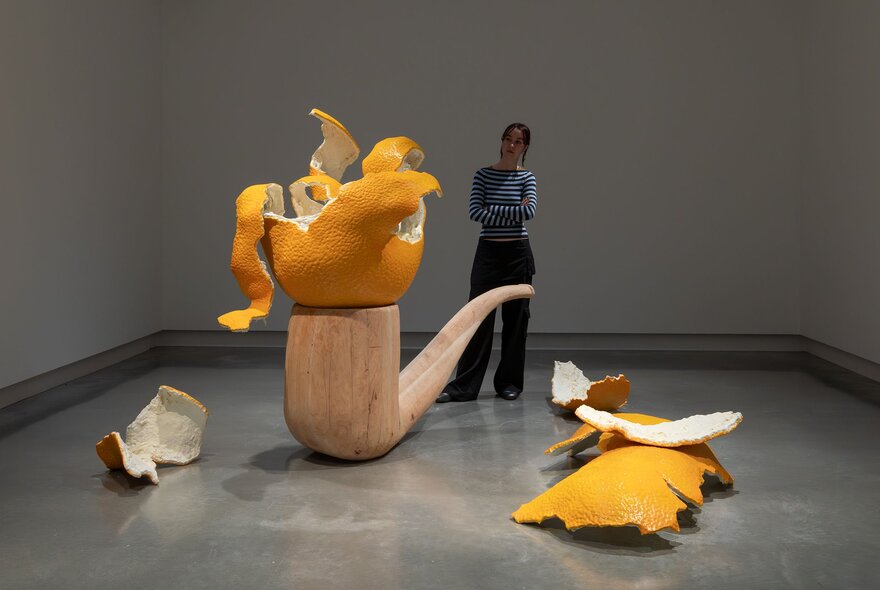 A person in a gallery setting looking at a sculpture of an oversize wooden pipe with a sculpture of a giant peeled mandarin balanced on the pipe.
