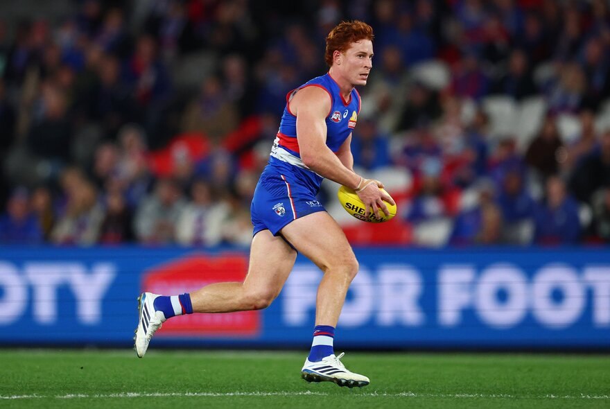 A Western Bulldogs AFL player running while holding a yellow football, with blurred fans in the stadium in the background.