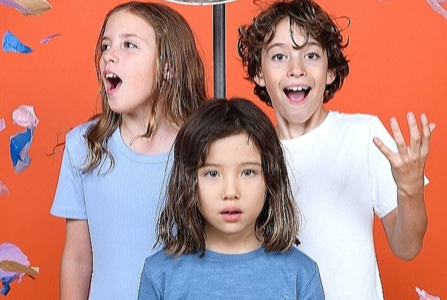 Three children, looking around in awe, in front of a red wall.