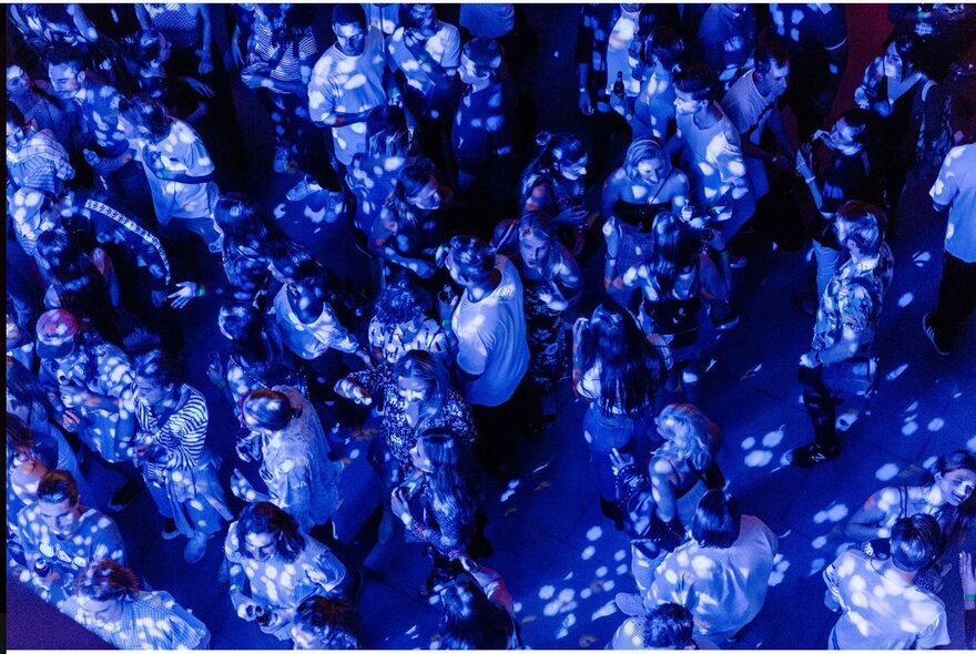 Patrons dancing on a blue-lit dancefloor at a nightclub, seen from above.