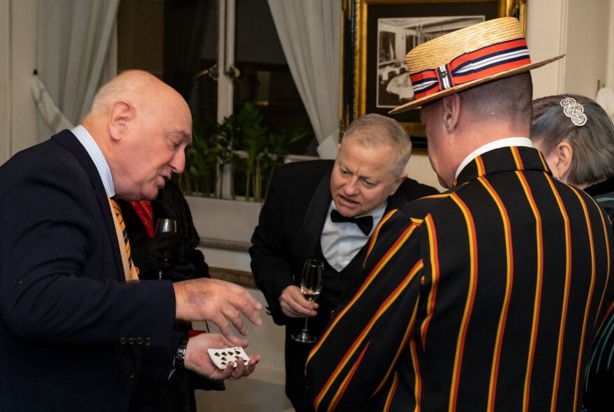 A man wearing a suit performing close up sleight of hand card tricks to a couple who watch on while holding drinks in thier hands inside a room.