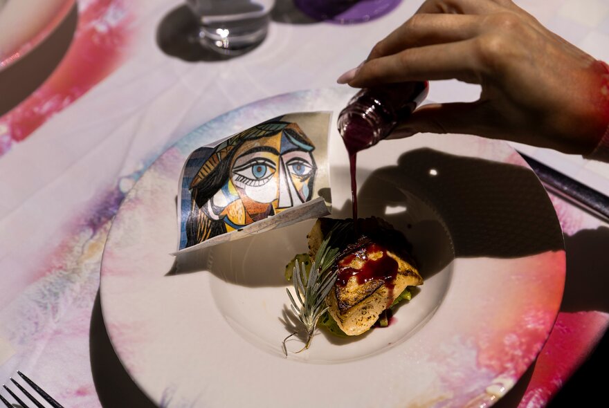 A hand pouring sauce from a small glass vial onto a fillet of fish on a plate, on which also rests a postcard image of a Picasso artwork.