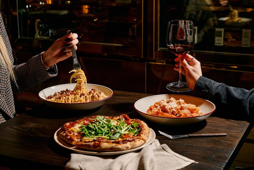 Hands twirling pasta and wine over dishes of pasta and pizza at a restaurant table.