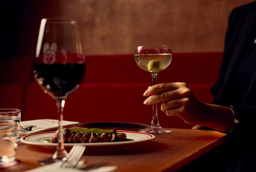 Restaurant table seating with plate of food, glass of wine and hand holding a martini with olive.