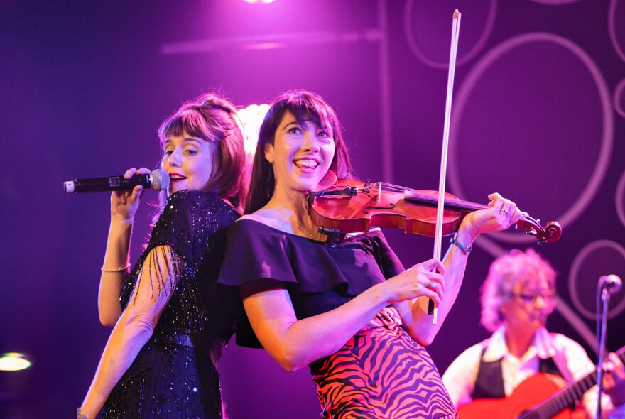 Two women back-to-back, one singing and one playing a violin and smiling broadly, a guitarist just visible in the background. 