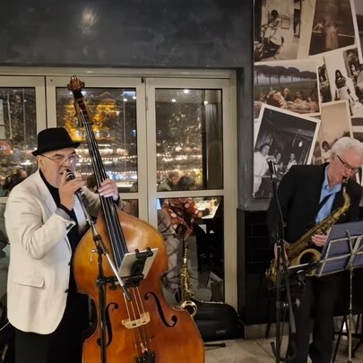 Two jazz musicians, one wearing a hat and playing an upright bass and one playing a saxophone in a small area in front of glass doors. 