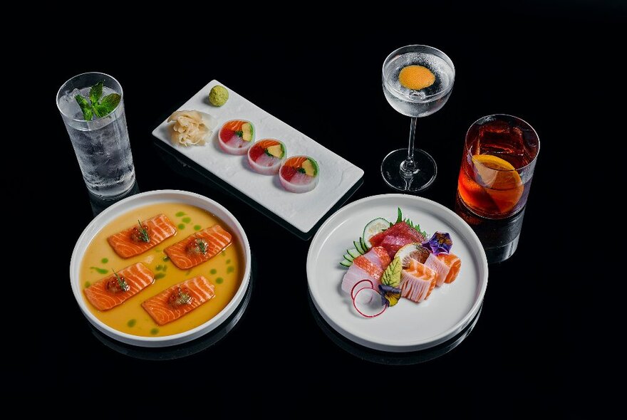 Three plates of elegantly presented sushi and sashimi, with three different drinks next to each dish, on a black background.