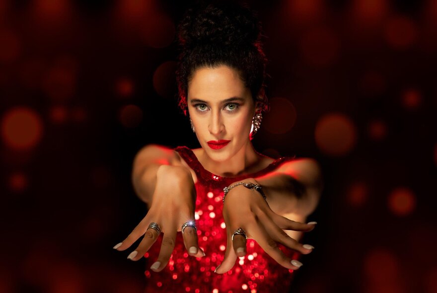 Vocalist Danielle Matthews wearing a red shimmering dress, with both her arms stretched out towards the viewer, flexing her fingers, against a shadowy background.