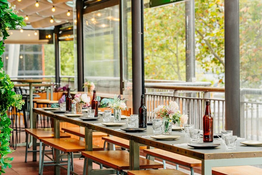 An empty restaurant, set for table service, with leafy green trees behind large glass windows.