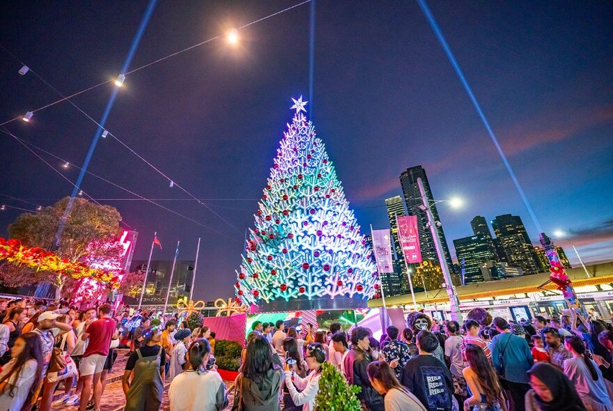 Crowds gathered around the Christmas tree at Fed Square at dusk, with city buildings in background.