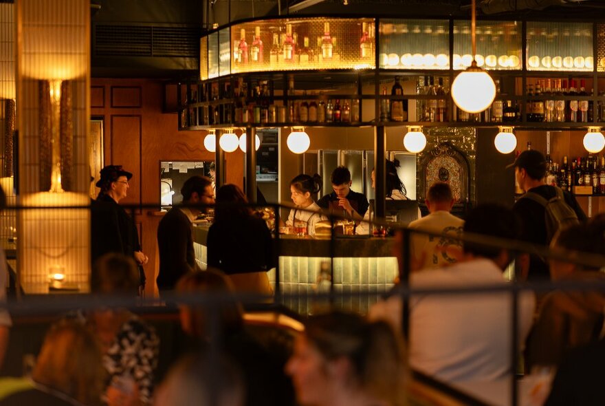 Interior of a venue with a rounded central bar, atmospheric round lights hanging from the ceiling, and people standing and sitting having drinks and snacks.