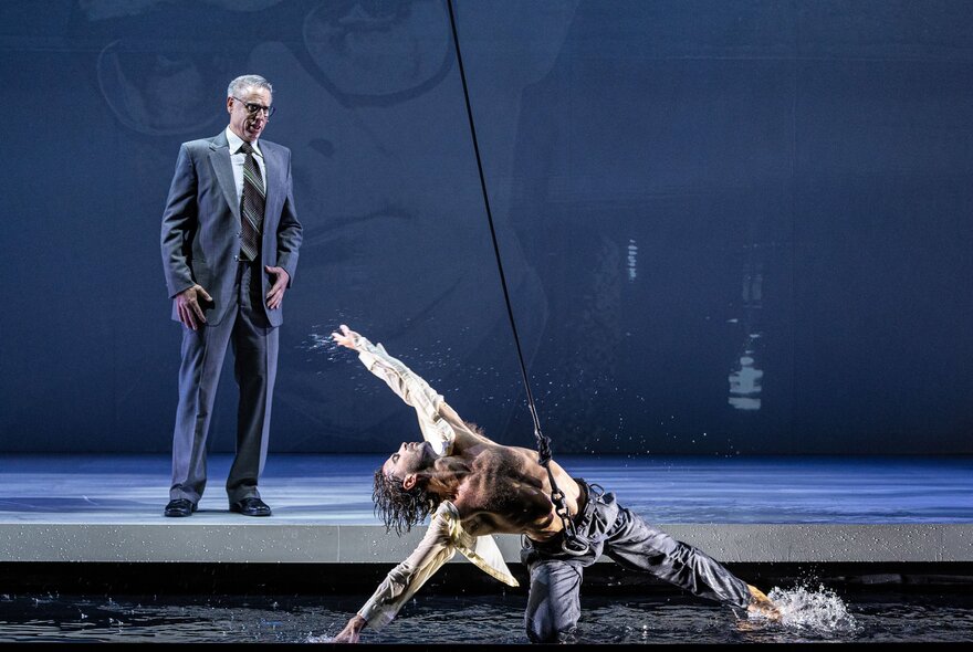 Two performers on a stage, a man dressed in a suit, looking at a man with his arms outstretched suspended by an aerial rope above a low pool of water.