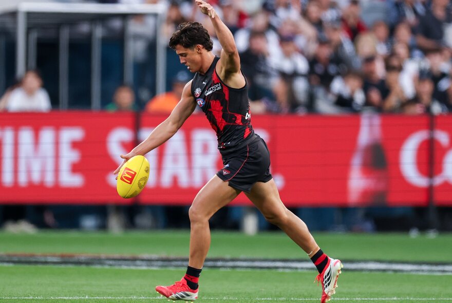 An Essendon AFL football player about to kick the ball during a match, the blurred stadium crowd in the background.