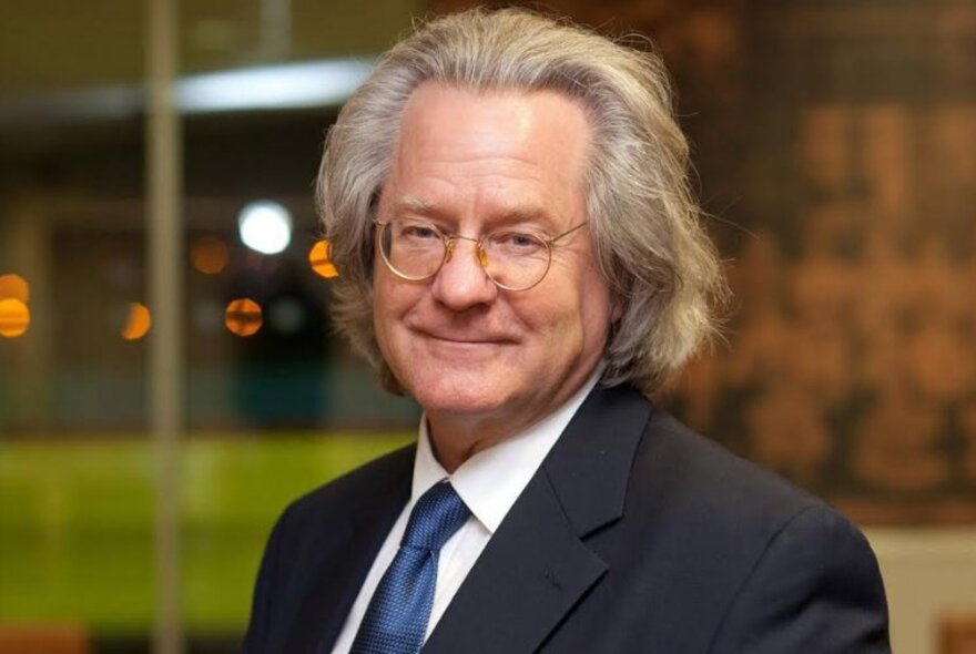 Portrait of preeminent philosopher, A. C. Grayling, wearing glasses, dark grey suit and a blue tie, subtly smiling at the camera.