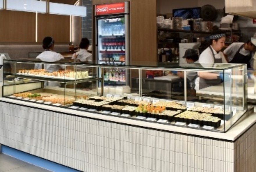 The food display counter at Maki Roll with sushi and maki rolls on trays behind glass, and staff in the open kitchen preparing food.