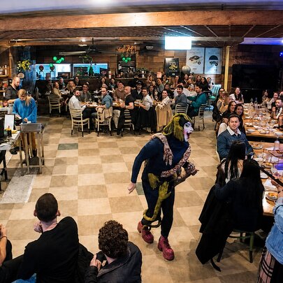 A drag queen performing in a restaurant setting while patrons are seated at long tables.