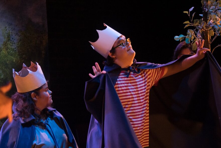 Two performers on stage, each wearing a cape and a paper crown, one with arms outstretched.