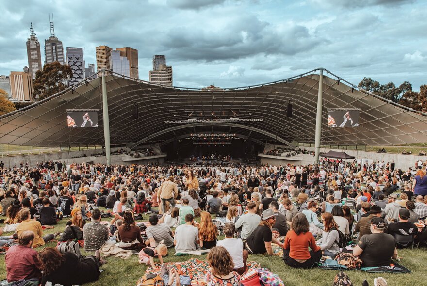 A crowd at the Sidney Myer Music Bowl in Melbourne, seen from the top of the hill, Melbourne's city buildings in the distance.