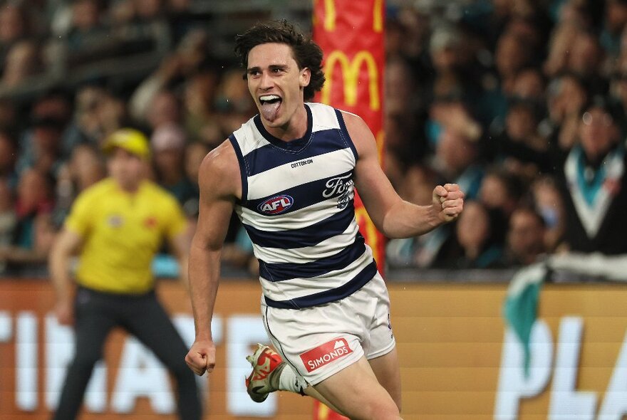 A Geelong player running after kicking a goal, his tongue out and making a fist, with an umpire visible in the background. 